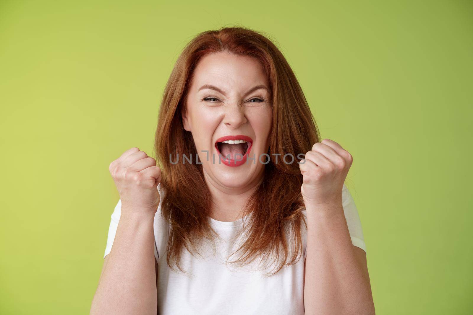 Yeah we did it. Joyful lucky redhead middle-aged female winner pump fists up celebration success gesture yelling triumph joy smiling broadly celebrate awesome news stand green background happy.