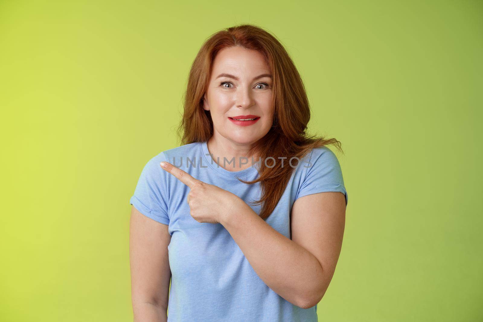 Surprised excited middle-aged wondered redhead woman pointing left amused standing thrilled joyful green background look camera curious interested cannot wait check-out great promo.