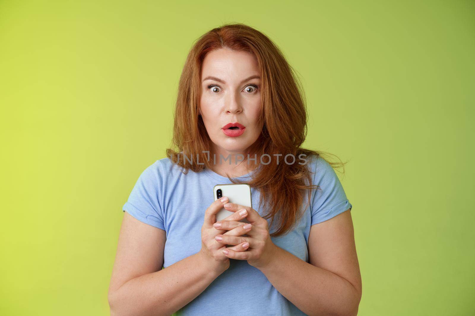 Shocked speechless impressed redhead european middle-aged woman fold lips astonished wow stare camera fascinated react amazed wondered received message hold smartphone intense green background.