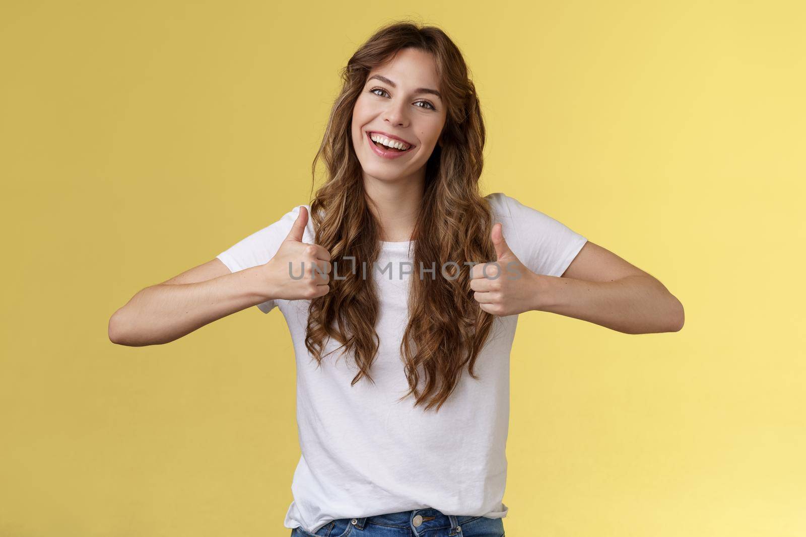 Outgoing positive confident lively young girl long curly haircut support great effor show thumbs up positive reply approving good choice like your outfit smiling broadly yellow background. Lifestyle.