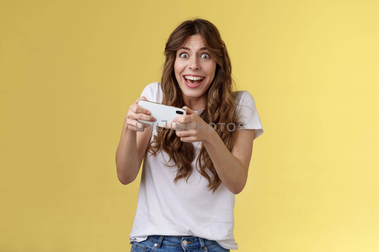 Happy enthusiastic excited cute playful girl winning awesome smartphone game thrilled playing stare camera surprised triumphing smiling happily hold mobile phone horizontal yellow background.