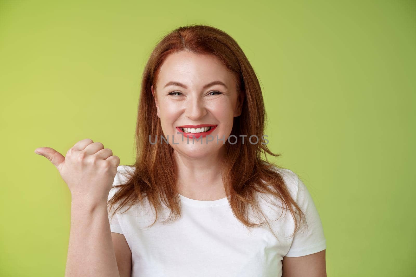 Come visit our store. Cheerful pleasant friendly charming redhead middle-aged woman entrepreneur inviting check-out promo smiling happily sincere kind grin pointing left thumb stand green background by Benzoix