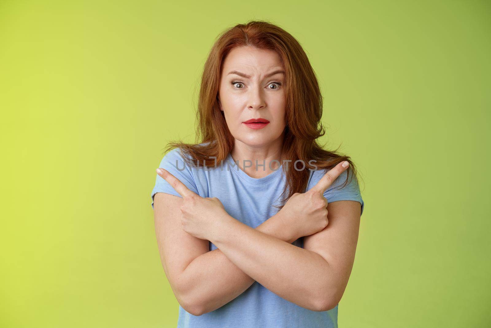 Help me choose. Worried unsure doubtful cute redhead middle-aged silly woman asking advice cross arms body pointing sideways left right look concerned camera make difficult decision green background by Benzoix