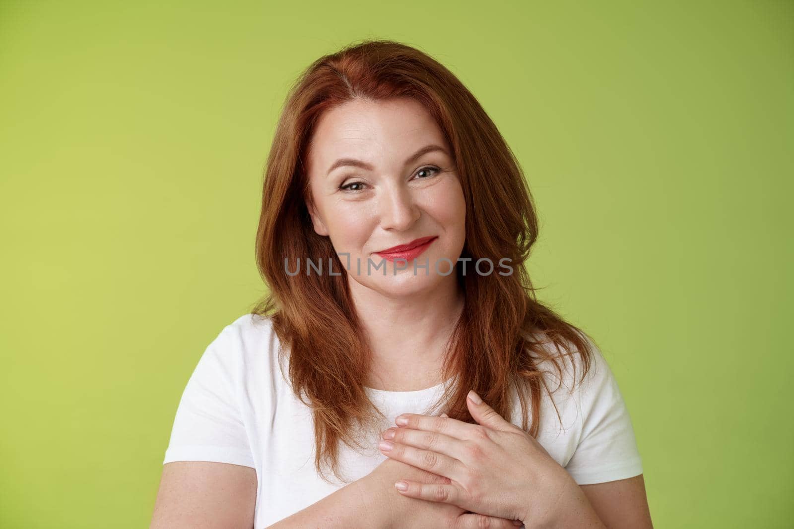 Lovely pleased tender redhead middle-aged ginger woman. press hands heart feel touched grateful appreciate sincere heartwarming moment admire thankfully grin look camera glad green background.