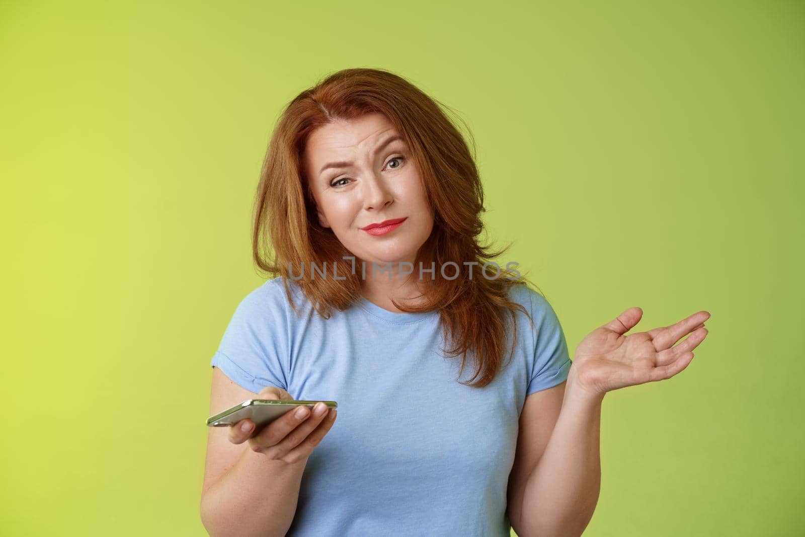 Well meh. Indifferent careless hesitant redhead middle-aged woman mature red female shrugging hold smartphone smirk bored uninterested hold hand aside apathetic attitude green background.