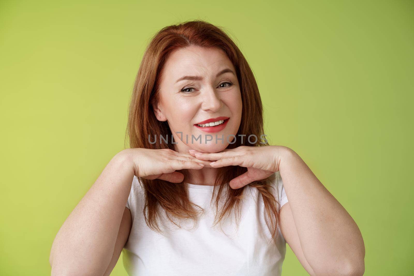 Looking good. Happy cheerful redhead middle-aged 50s woman smiling delighted hold hands under chin accept flaws blemished like own skin condition apply aging creme cosmetics green background.