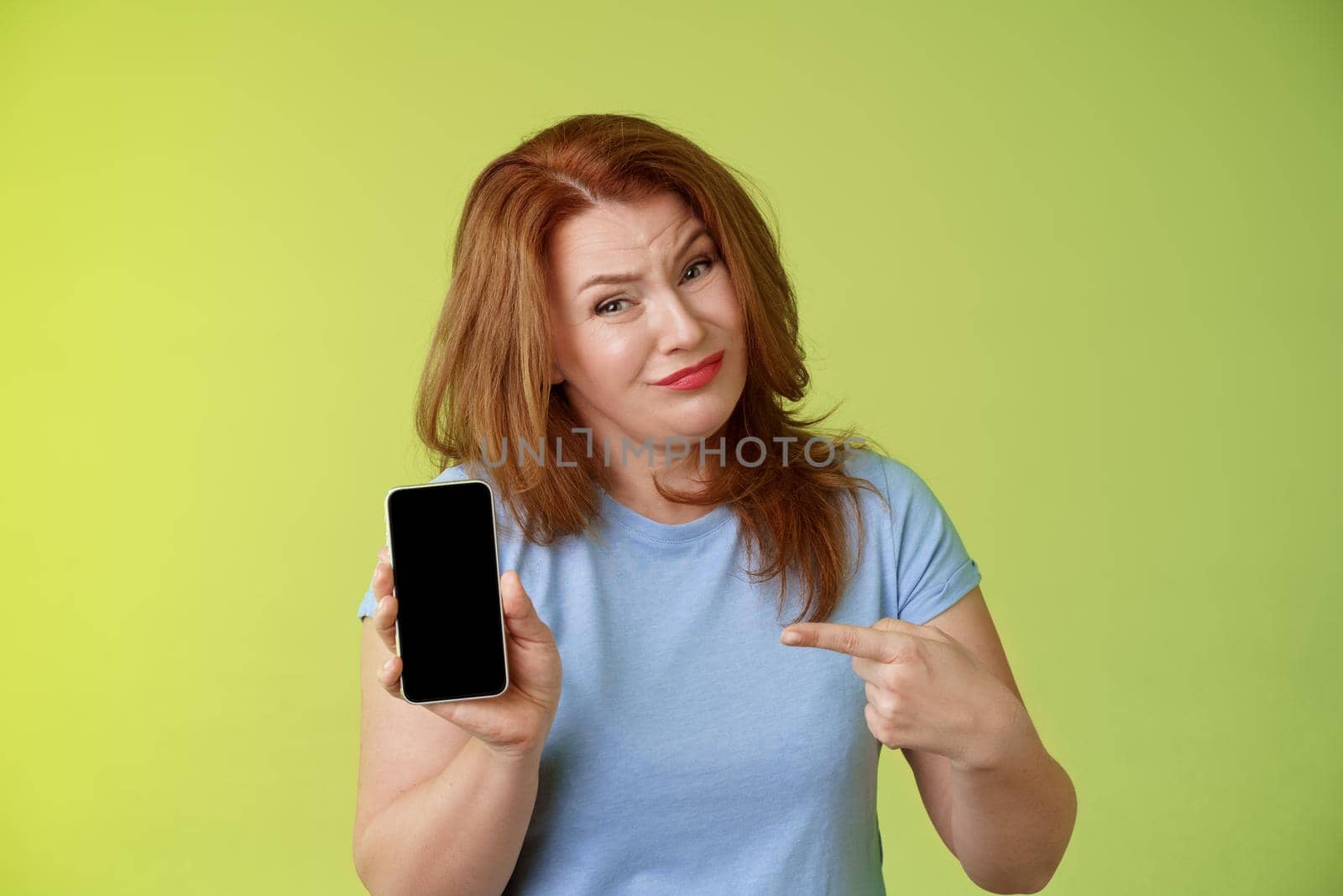 Seriously it awful. Displeased disappointed redhead mature female tilt head cringe grimacing reluctant pointing smartphone blank display index finger showing bad photograph share negative opinion.