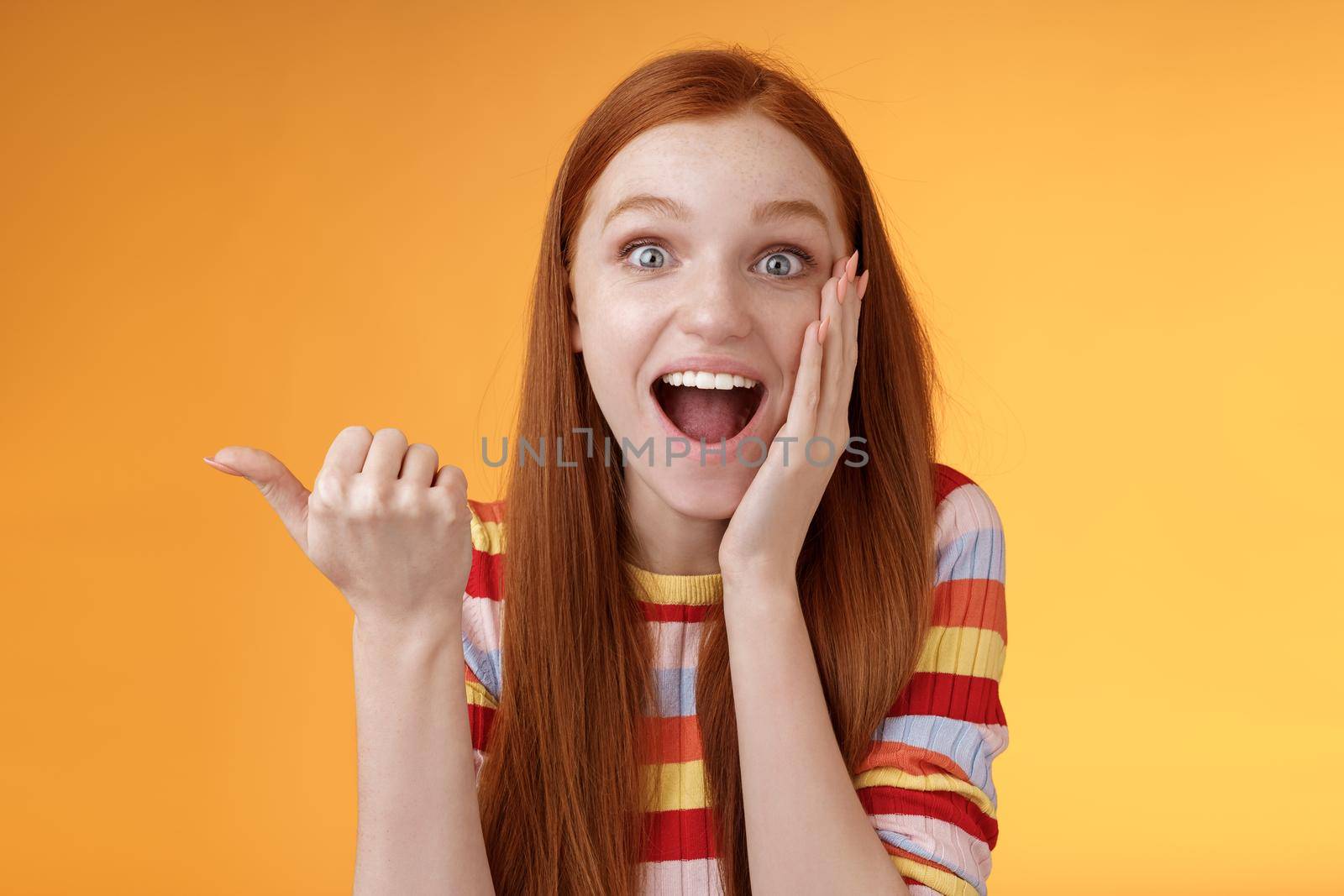 Cheerful excited impressed talkative girl pointing astonished thumb left drop jaw smiling broadly touch cheek thrilled, interesting event discuss awesome wonderful performance, orange background.