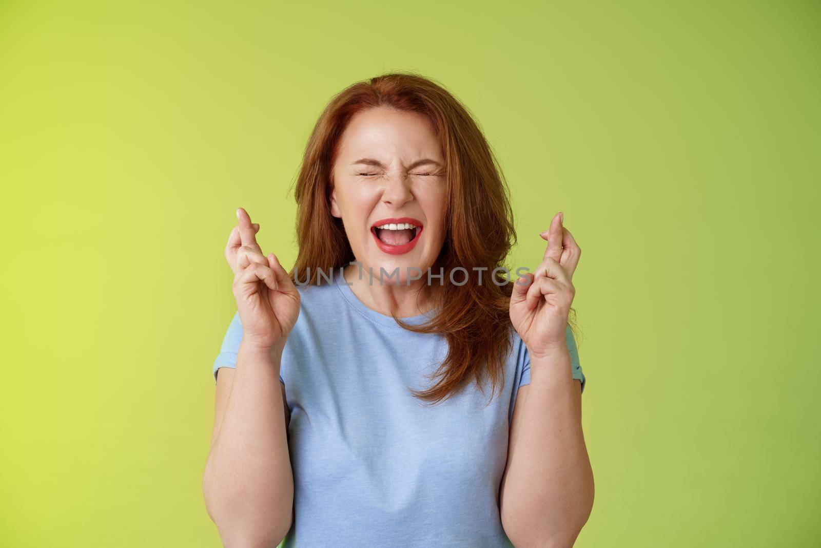 Woman wants win badly. Enthusiastic lucky redhead middle-aged 50s female pleading implore god make dream come true cross fingers good luck wishing closed eyes open mouth excitement green background by Benzoix