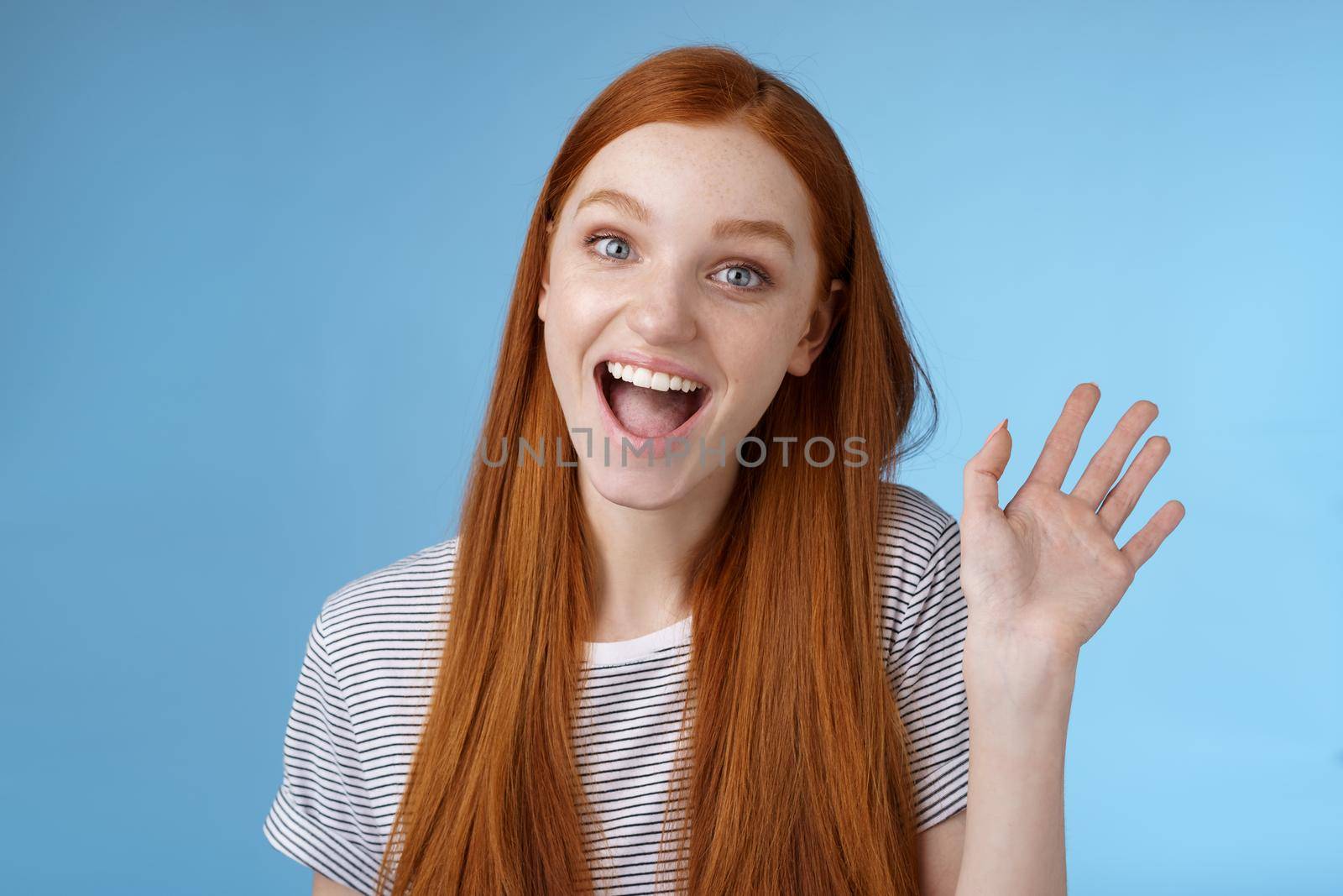 Hello wanna be friends. Enthusiastic cute redhead female newbie getting know coworkers smiling happy waving raised hand hi greeting gesture welcoming, say bye standing blue background by Benzoix