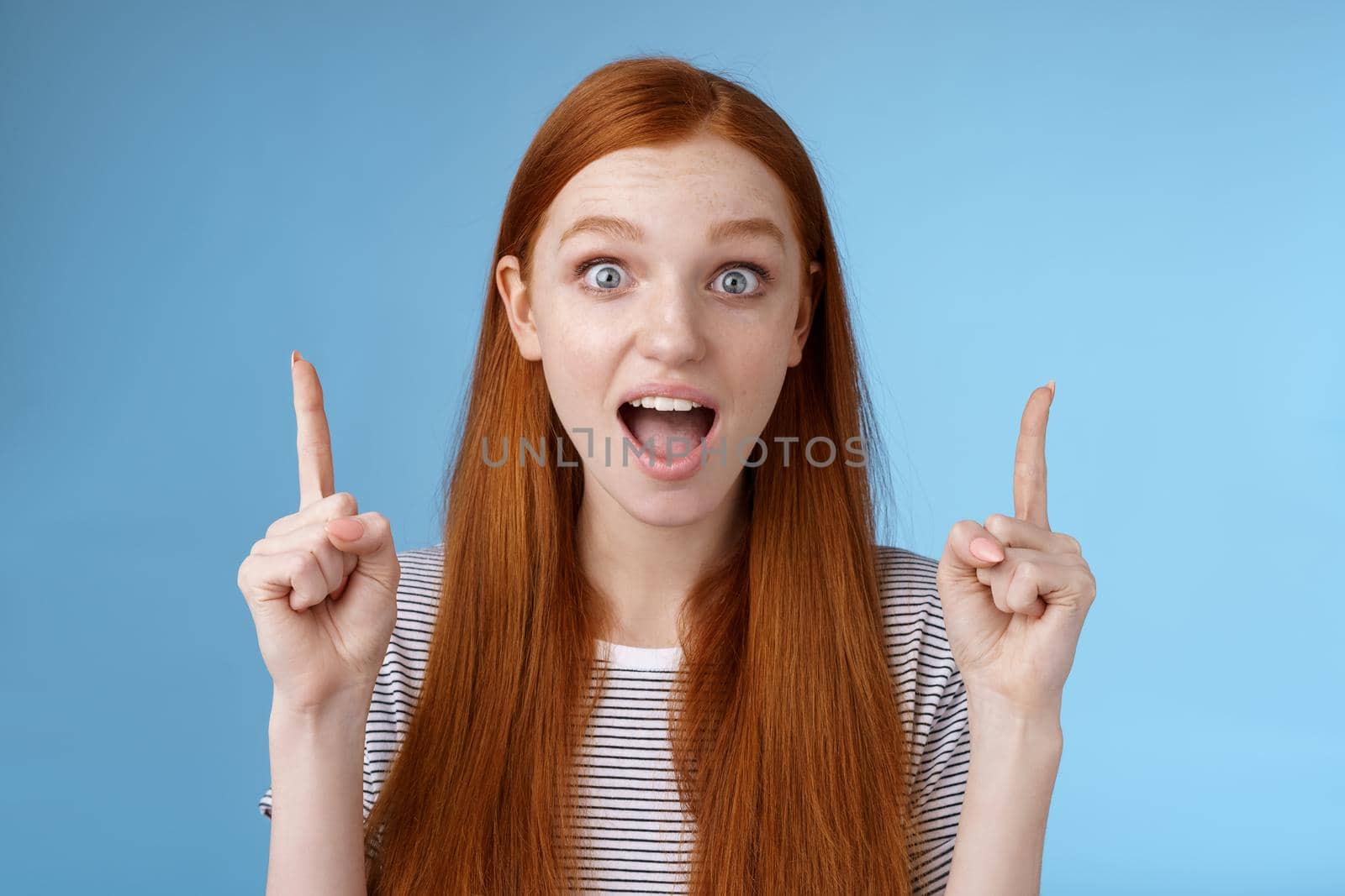 Surprised happy enthusiastic young redhead woman reacting impressed upper hanging promo pointing up index fingers drop jaw amused look thrilled camera telling about awesome offer, blue background by Benzoix