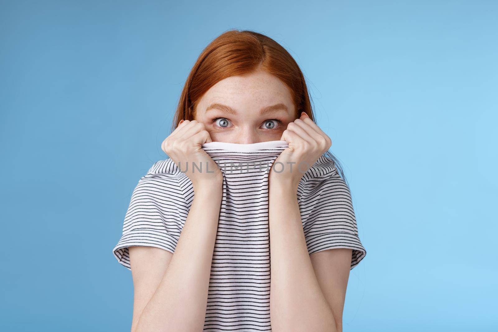 You cannot see me. Silly cute young teenage redhead girl hiding face pulling t-shirt head peeking look excited surprised camera hold collar standing funny under disguise, blue background by Benzoix