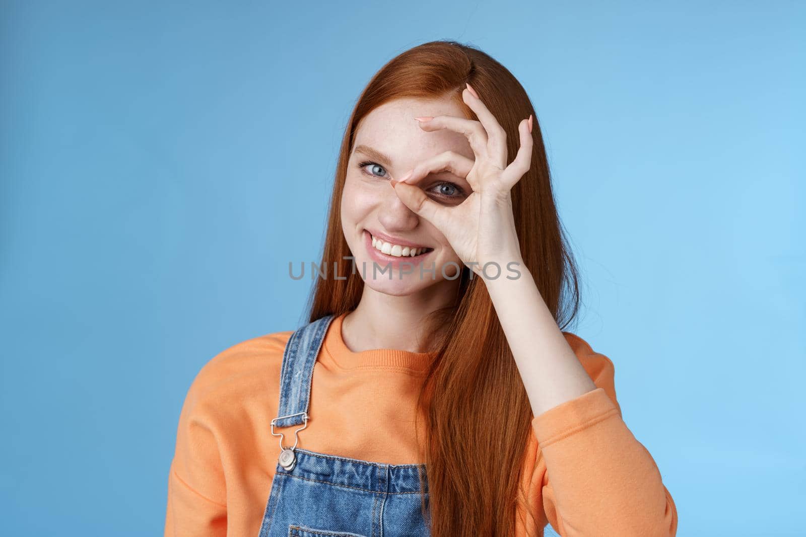 Girl search perfection. Charming pretty glad redhead female student assuring everything ok showing okay sign eye look through smiling delighted express positive attitude, blue background by Benzoix