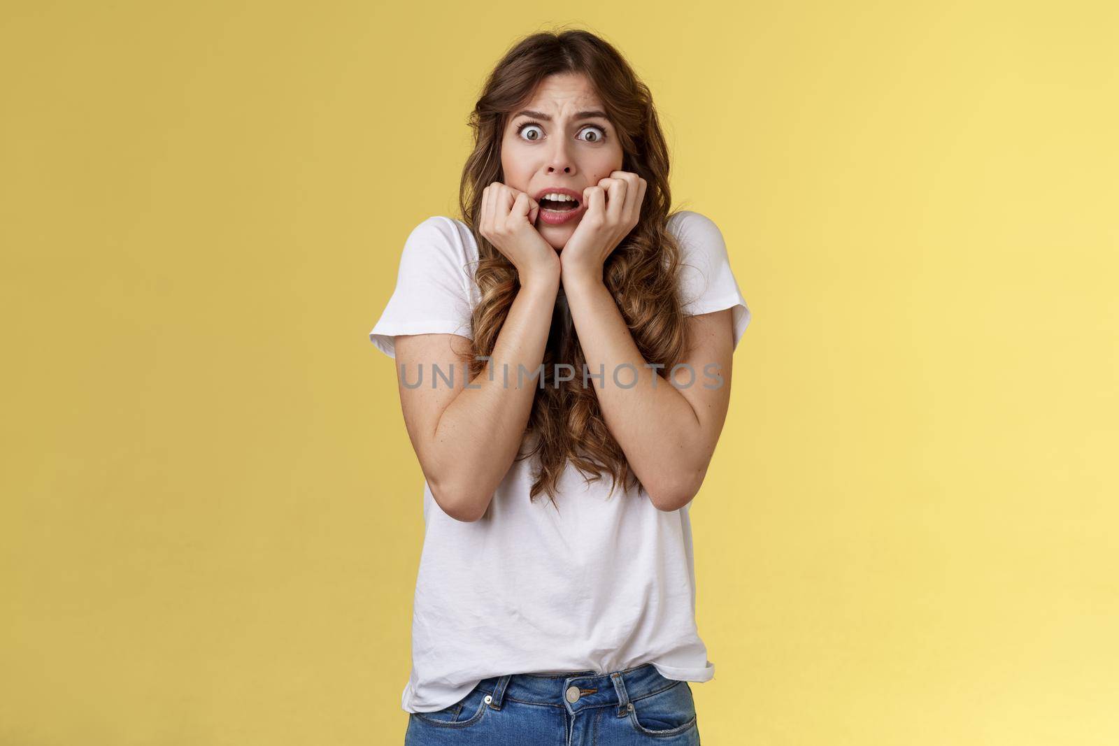 Timid insecure intense gasping young scared attractive woman sighing shocked frightened hold hands face biting nails terrified express fear standing shook yellow background stare camera.