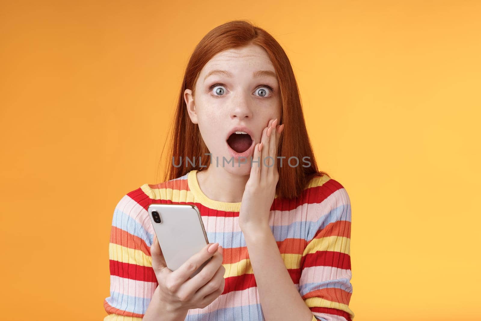 Concerned shocked emotive redhead girl find out stunning news look camera drop jaw gasping impressed touch cheek hold smartphone finish reading impressive disturbing story, orange background.