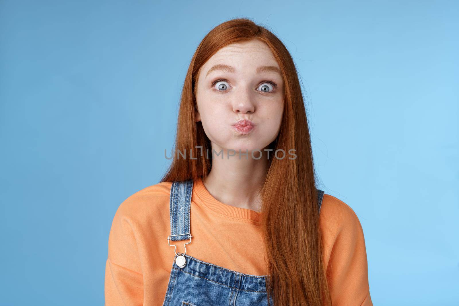 Cute childish playful charming redhead girl popping blue eyes staring camera hold breath inhale air mouth standing speechless promise not slip secret acting immature standing funny happily by Benzoix