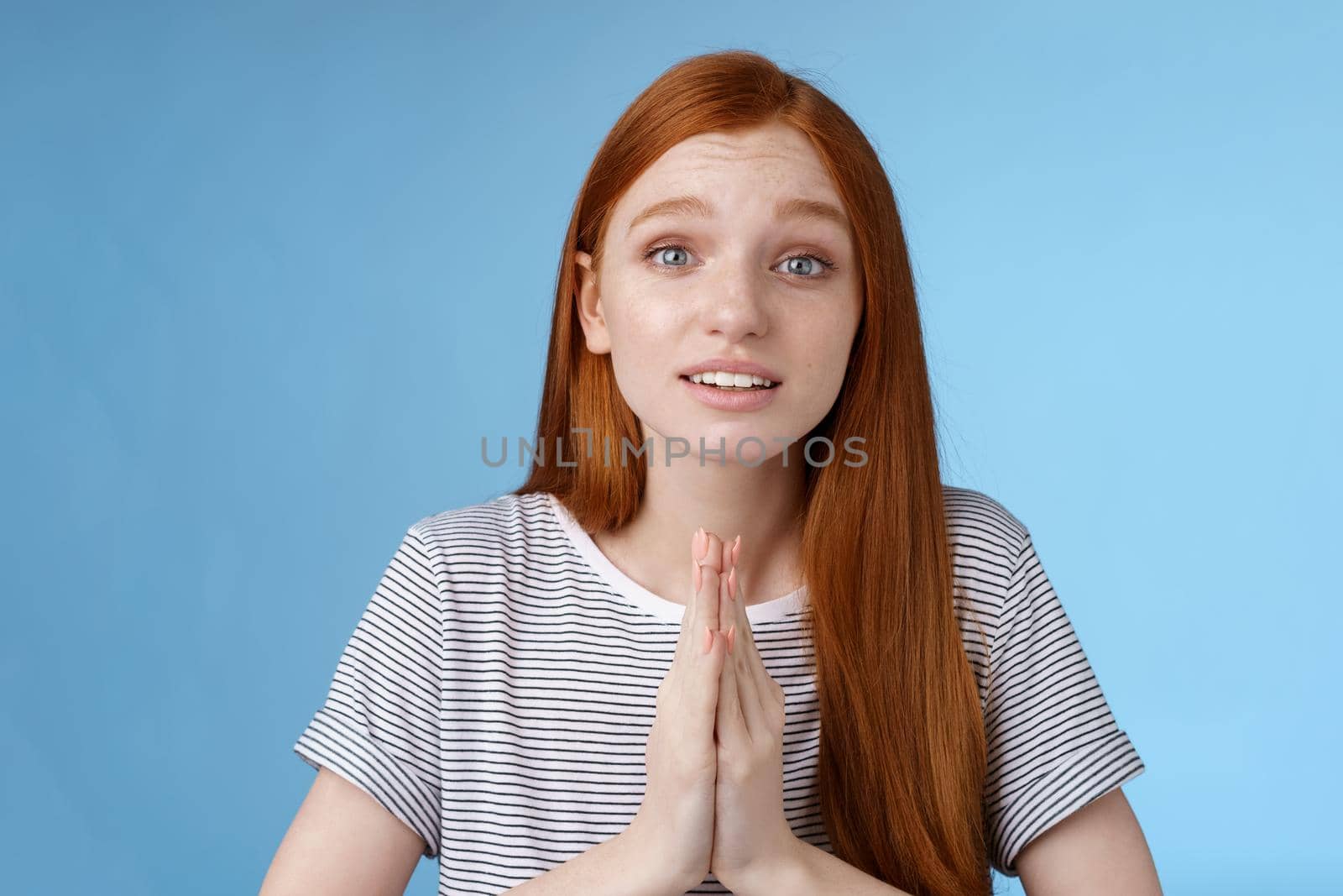 Cute european redhead girl blue eyes straight long ginger hairstyle making promise begging you help press palms together praying say please telling need favour worried pleading, blue background by Benzoix