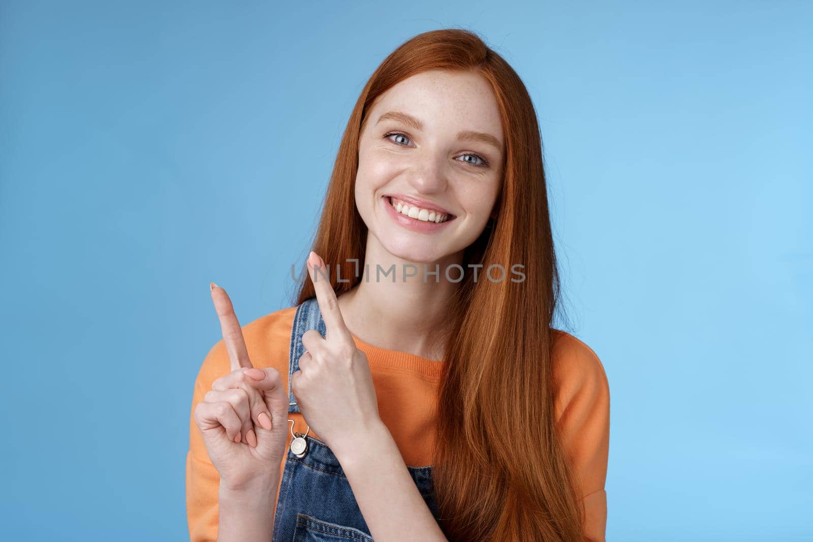 Lifestyle. Happy tender redhead girl sincerely smiling white teeth helpful look camera excited give hand pointing upper left corner introduce sale offer recommend try promo standing blue background.