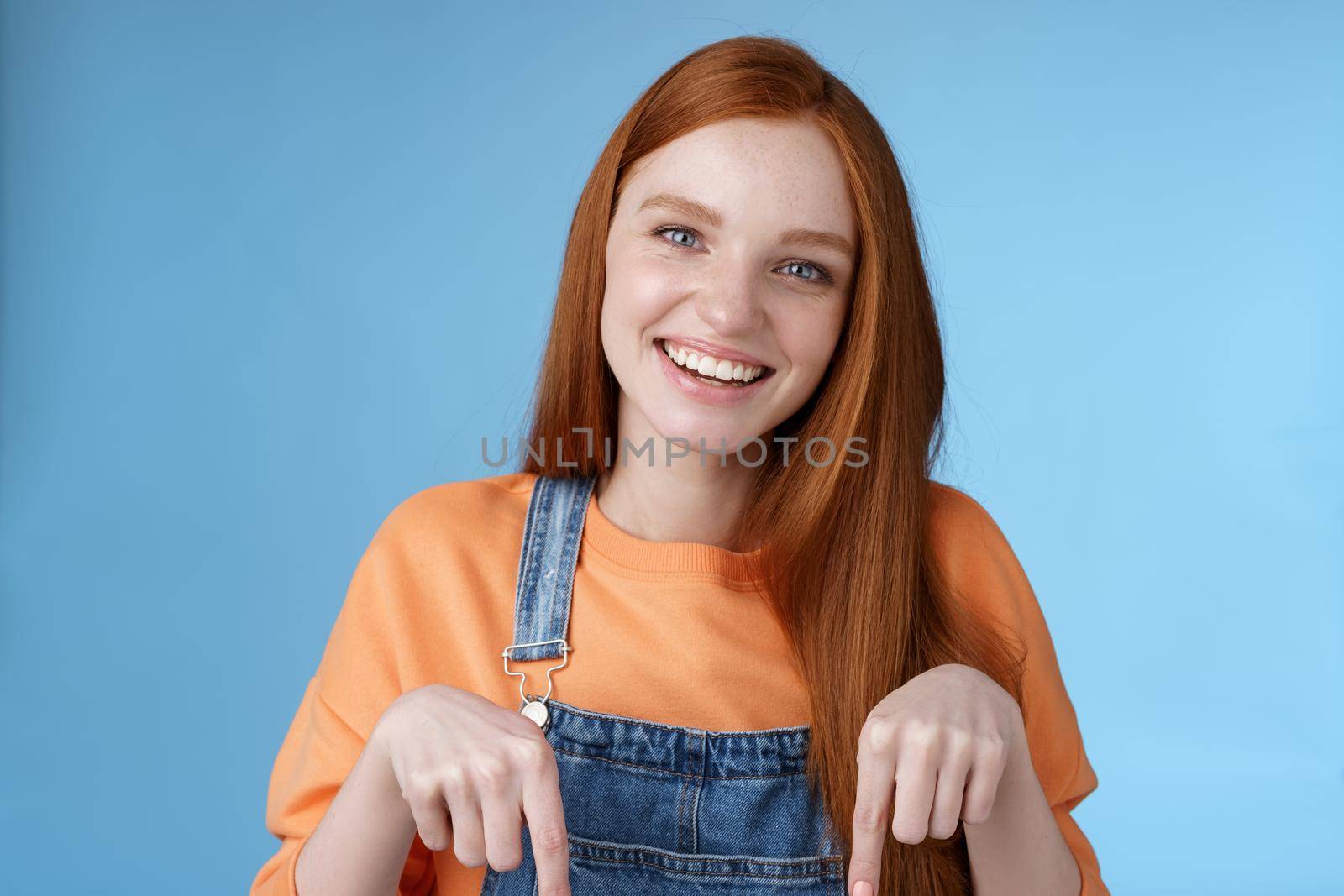 Friendly happy redhead girl smiling lively pretty grin pointing down index fingers offering good offer recommend use service standing blue background discuss interesting product, blue background by Benzoix