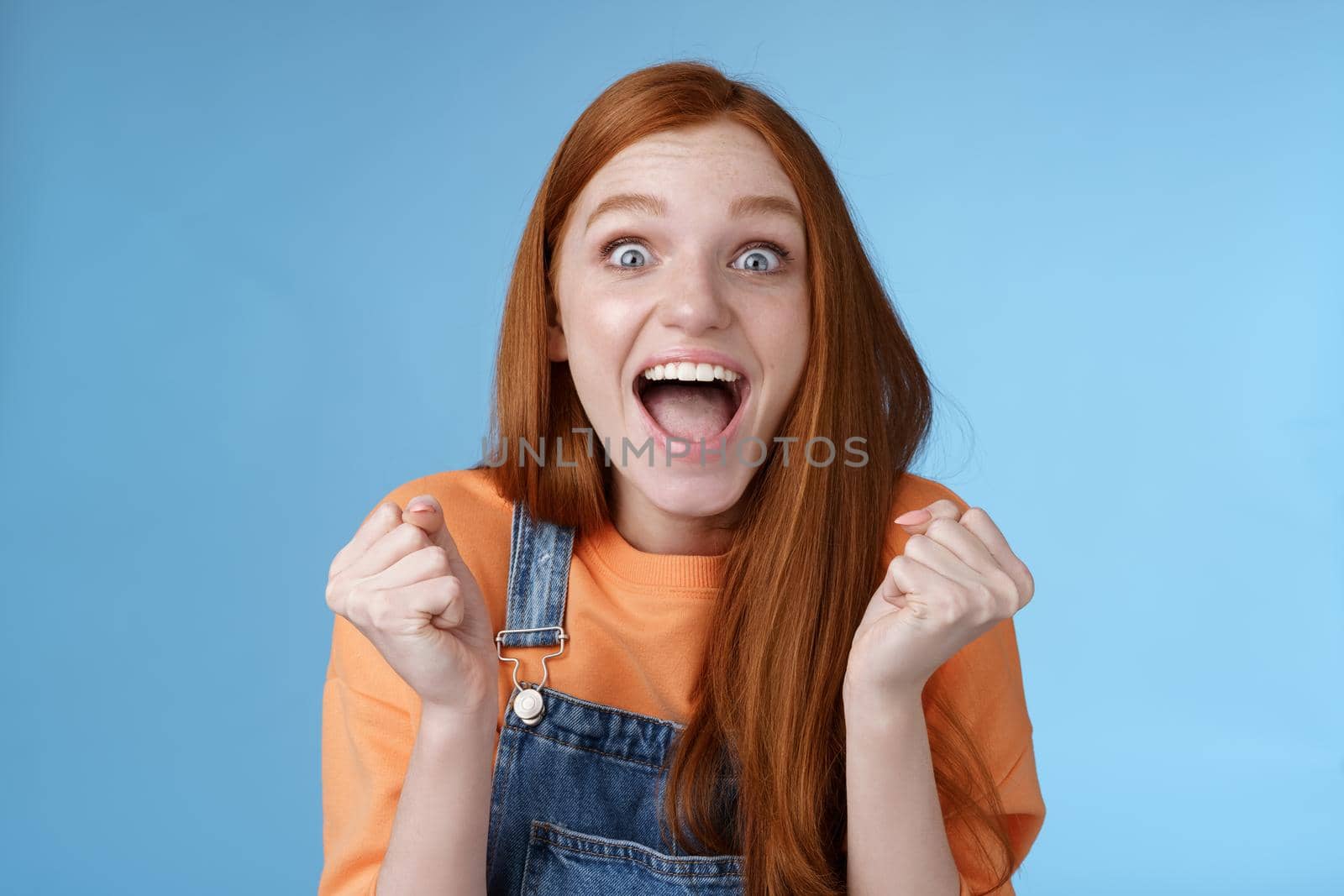 Excited happy overwhelmed cute ginger girl blue eyes yelling out loud rejoicing fantastic awesome news clench fists triumphing celebrating victory win standing amazed blue background achieve success by Benzoix