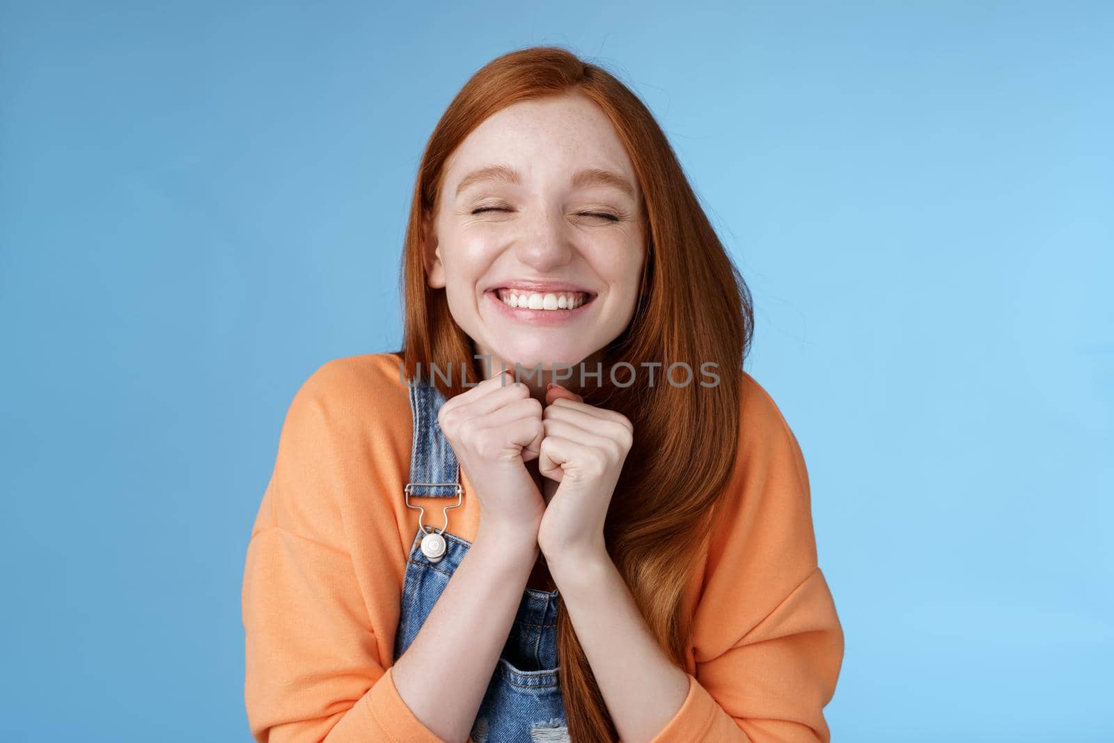 Eager rejoicing thrilled pretty young redhead girl close eyes dreamy smiling receive great result scholarchip triumphing joyfully grinning squeez hands excited, standing blue background very happy by Benzoix