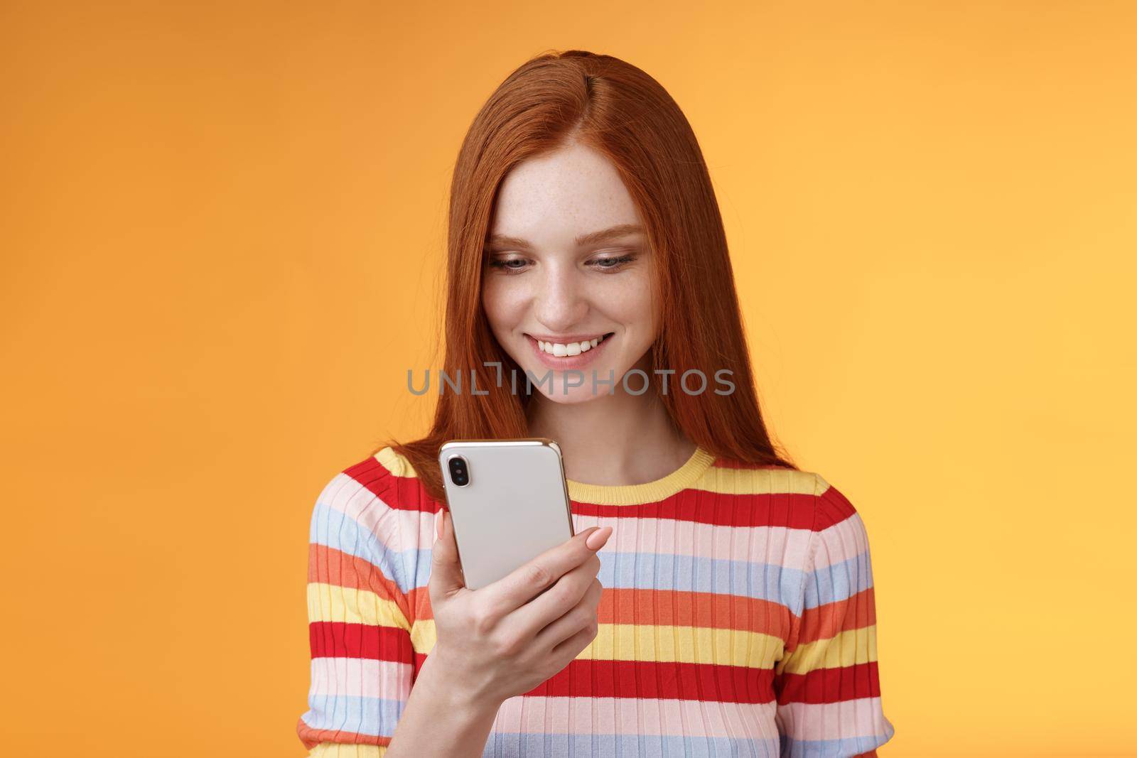 Charming modern redhead girl college student checking message box holding smartphone look happy smiling delighted cellphone display receive hundred likes photo post online, orange background.