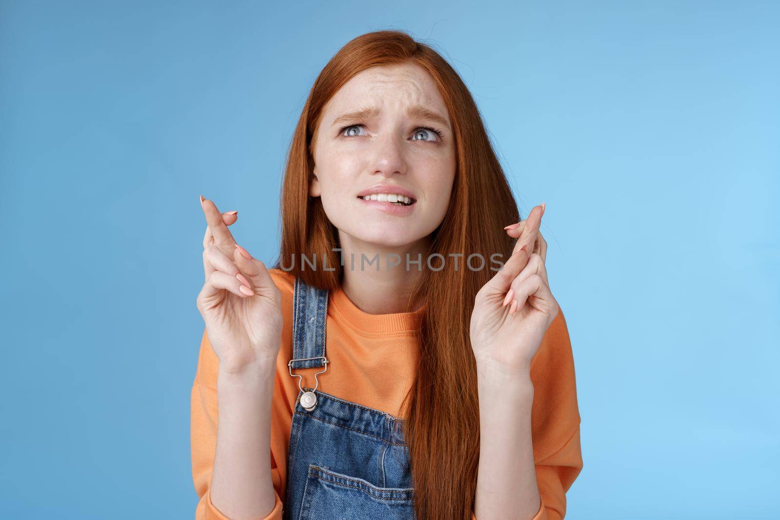 Worried unconfident young redhead intense cute girl frowning nervously waiting improtant moment cross fingers good luck bite lip anxiously frowning look upper left corner praying wish come true by Benzoix