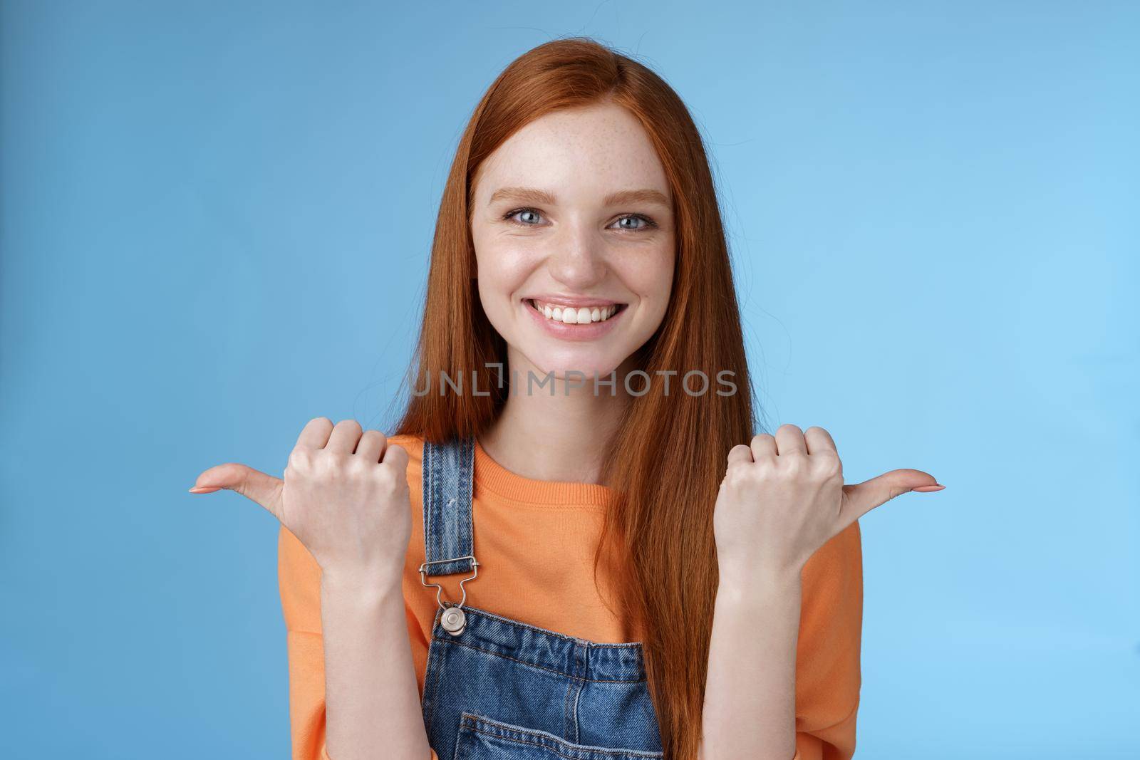 Indoor shot charismatic assertive happy smiling redhead woman orange shirt denim overalls pointing sideways thumbs left right showing choices opportunities give chance choose, blue background by Benzoix