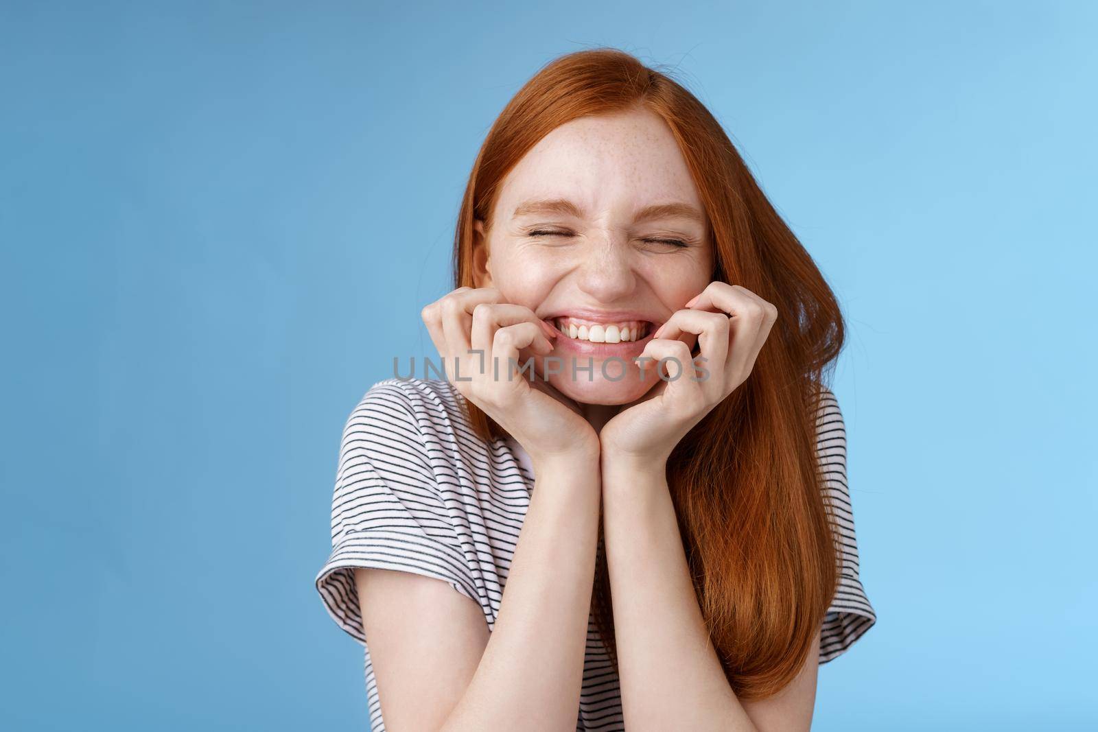 Cheerful carefree giggling ginger girl look happy bright close eyes smiling delighted hold hands cheeks having fun feeling excitement joy triumphing cheering good news, standing blue background by Benzoix
