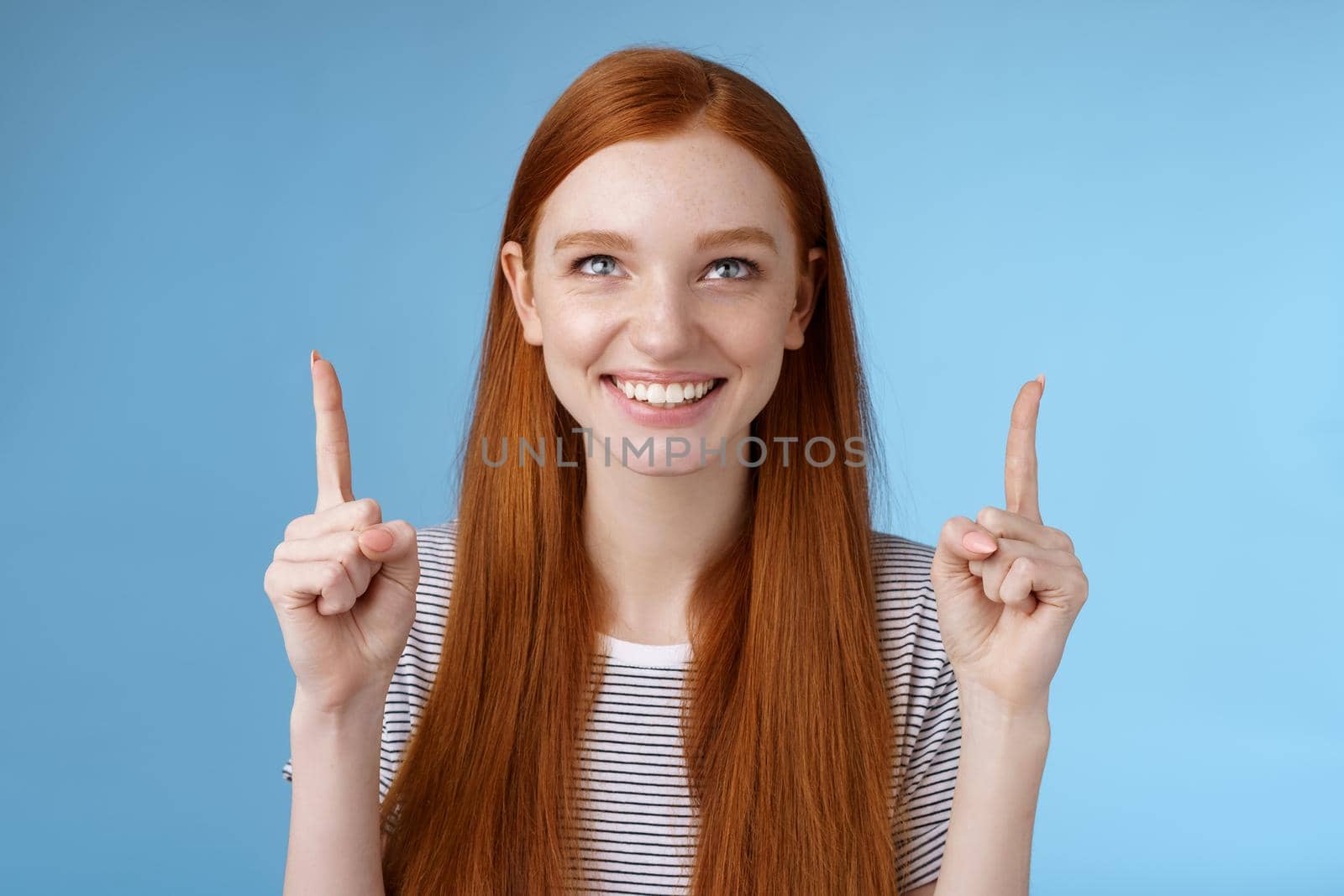 Amused excited happy smiling redhead girl look pointing up grinning gladly watching awesome open air perfomance standing joyfully showing cool thing indicating product, blue background.