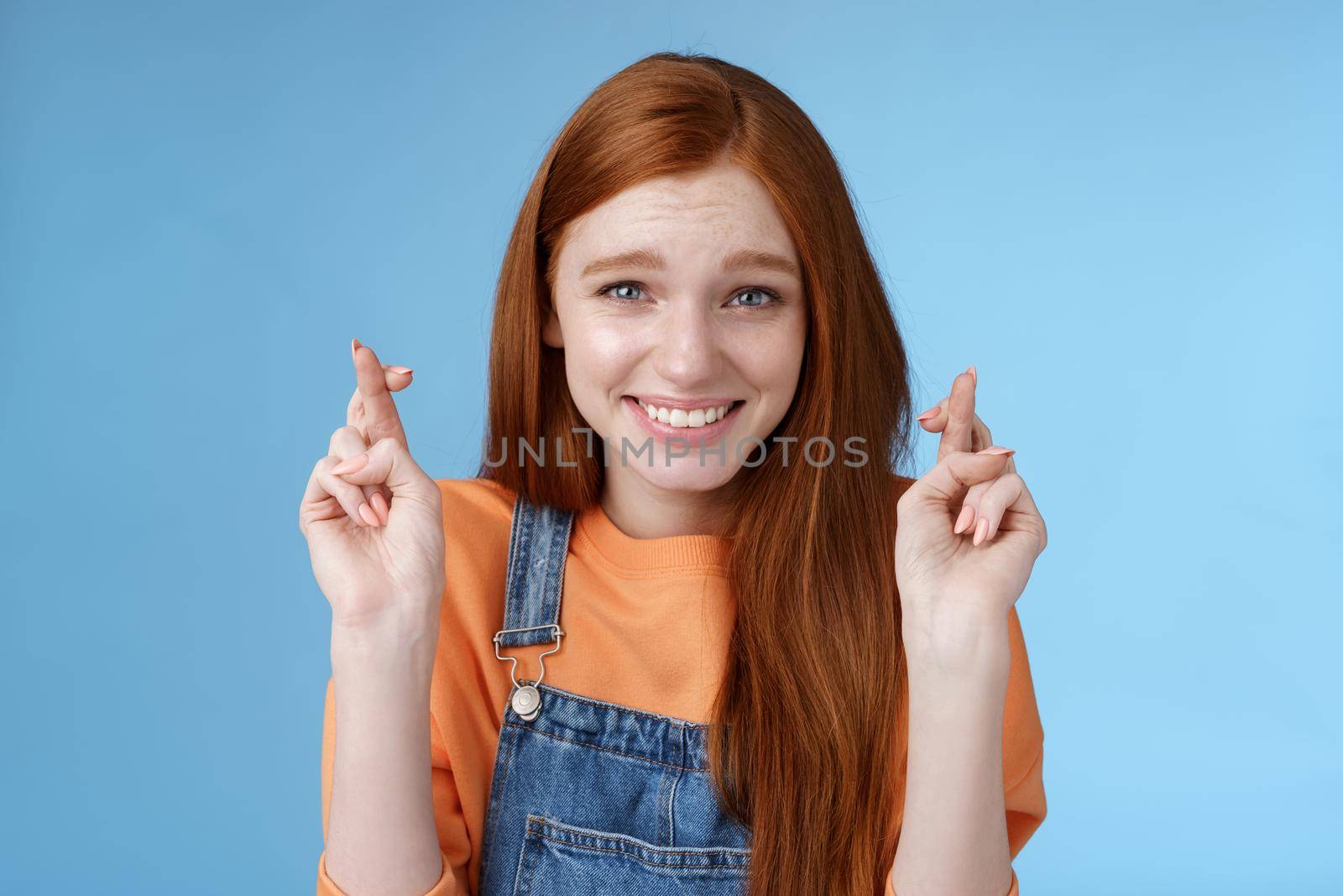 Lifestyle. Silly young pretty redhead girl blue eyes freckles making wish cross fingers good luck smiling hopeful eager get best results waiting desires fulfill miracle happen anticipating blue background.