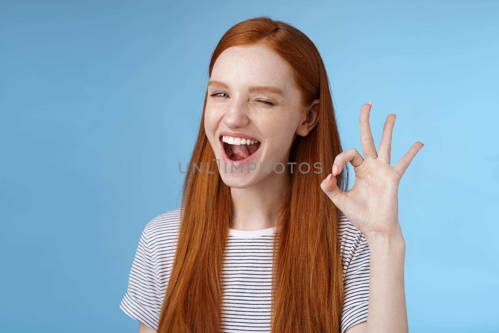 Sassy attractive redhead girl winking mysteriously smiling broadly give approval sign show okay ok excellent gesture satisfied good choice agree great decision, standing delighted blue background.