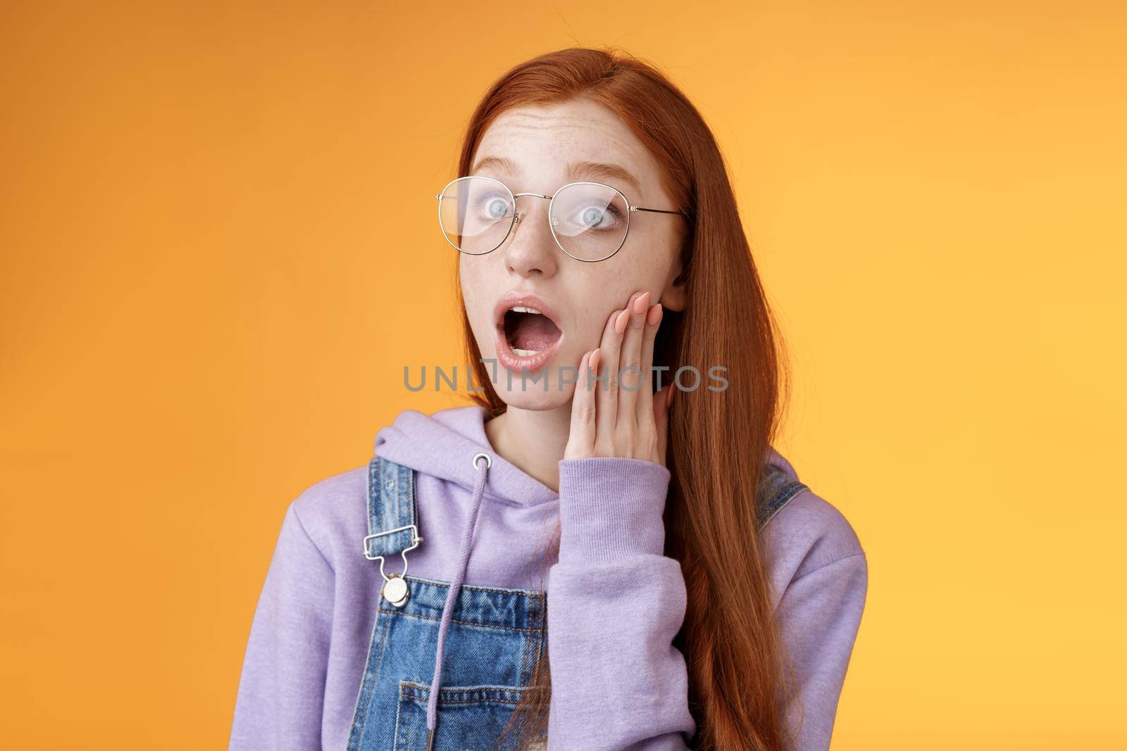Shocked impressed speechless young redhead sensitive european girl glasses wearing hoodie dungarees drop jaw gasping astonished look left wide eyes amazed standing orange background by Benzoix