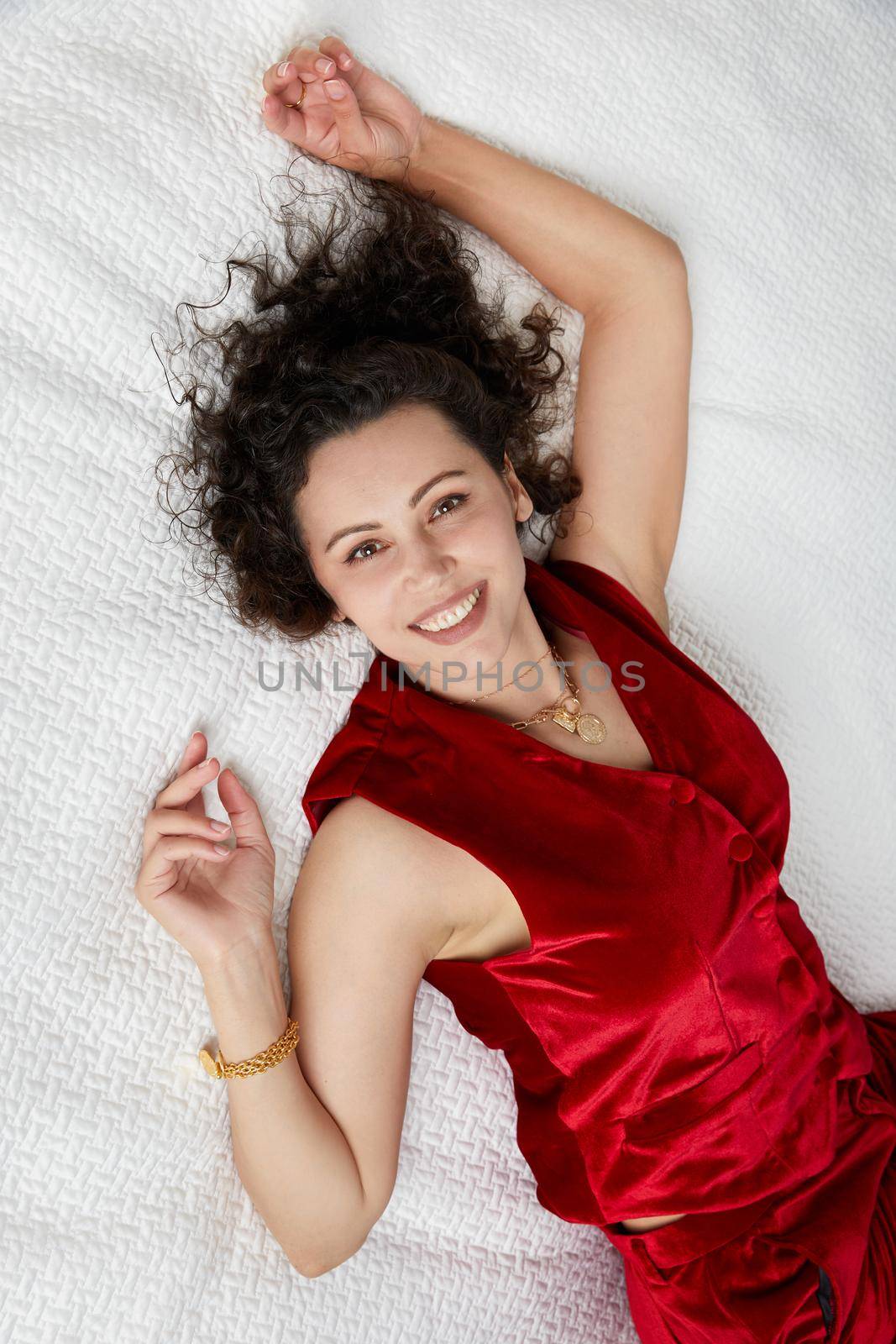 Top view of beautiful curly young woman wearing red velvet suit lying on bed and looking at camera