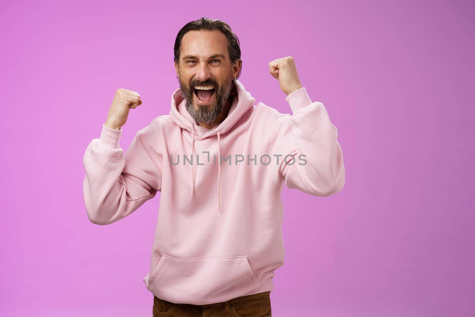 Cheerful supportive manly mature adult bearded guy fan yelling raising clenched fists triumphing team scored goal celebrating standing pleased shouting achieving success, posing purple background.