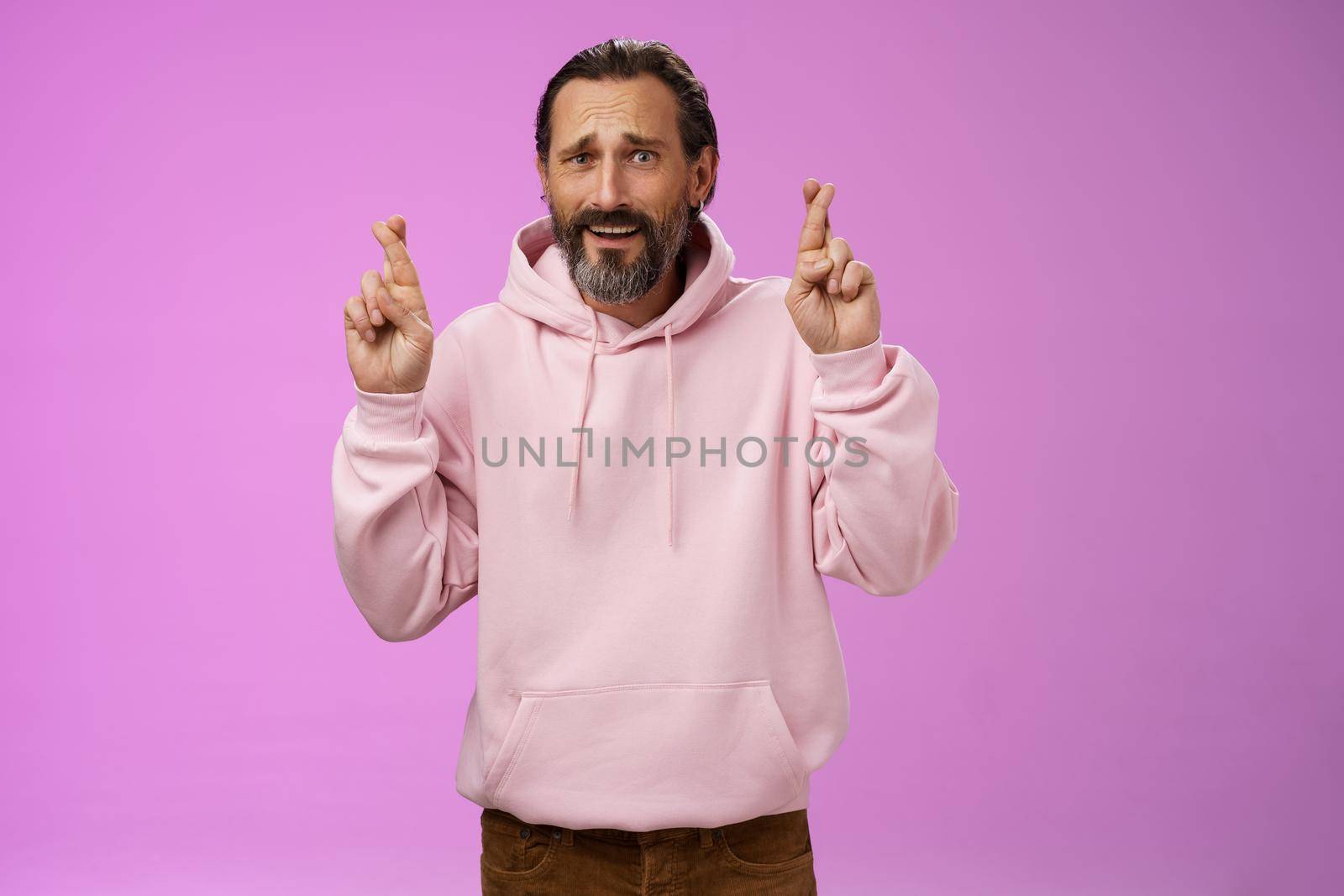 Nervous unsure hopeful handsome bearded 40s man in pink stylish hoodie cringing worried cross figers good luck make wish hope not gonna lose job supplicating praying dream come true. Lifestyle.