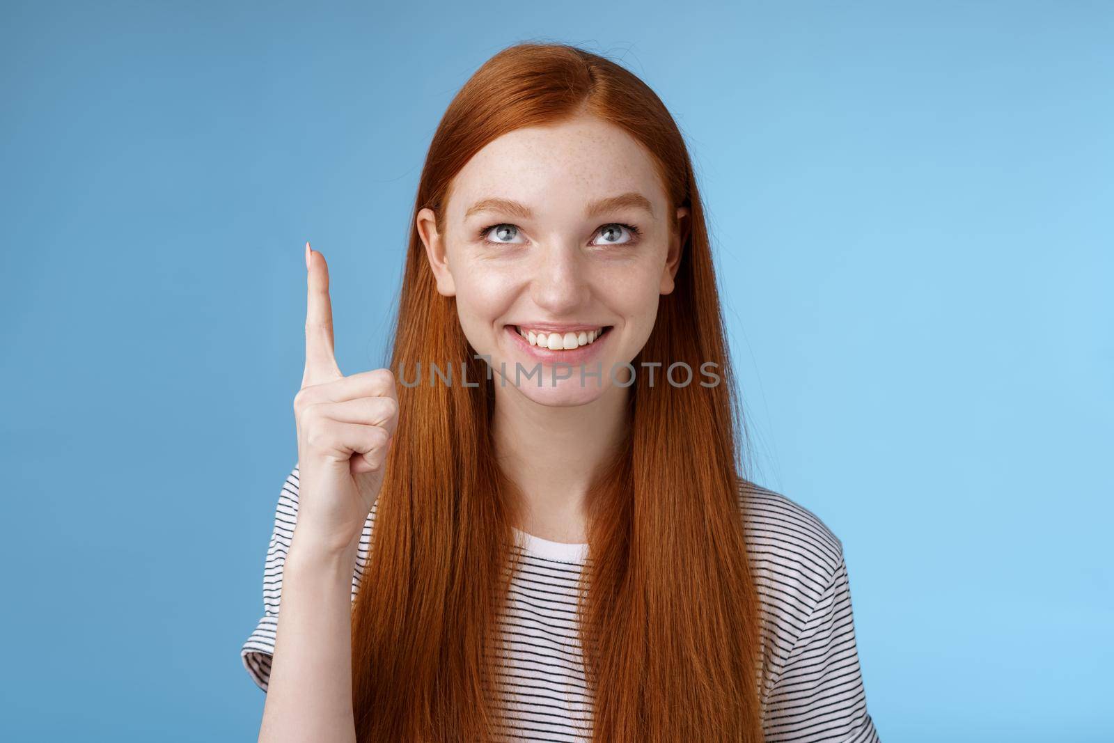 Intrigued lively happy smiling redhead european girl 20s look pointing up index fingers amused check out interesting sale holiday promo offer standing intrigued thrilled blue background. Lifestyle.