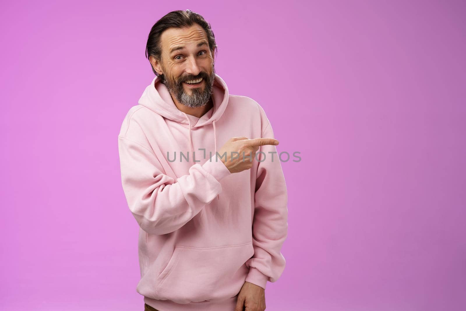 Friendly carefree caucasian bearded man in pink hoodie pointing right intoducing family members smiling proudly delighted showing blank copy space advertising, posing purple background.