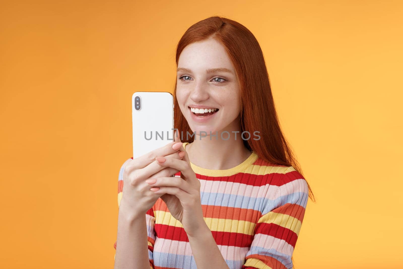Redhead girl having fun recording hilarious friend actions hold smartphone look display amused shooting funny video telephone standing orange background satisfied smiling delighted. Lifestyle