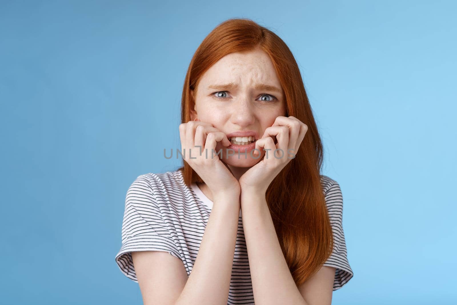 Worried uncomfortable scared young panicking redhead young girl feeling pressure distressed frowning squinting frightened biting fingernails trembling fear, standing blue background.