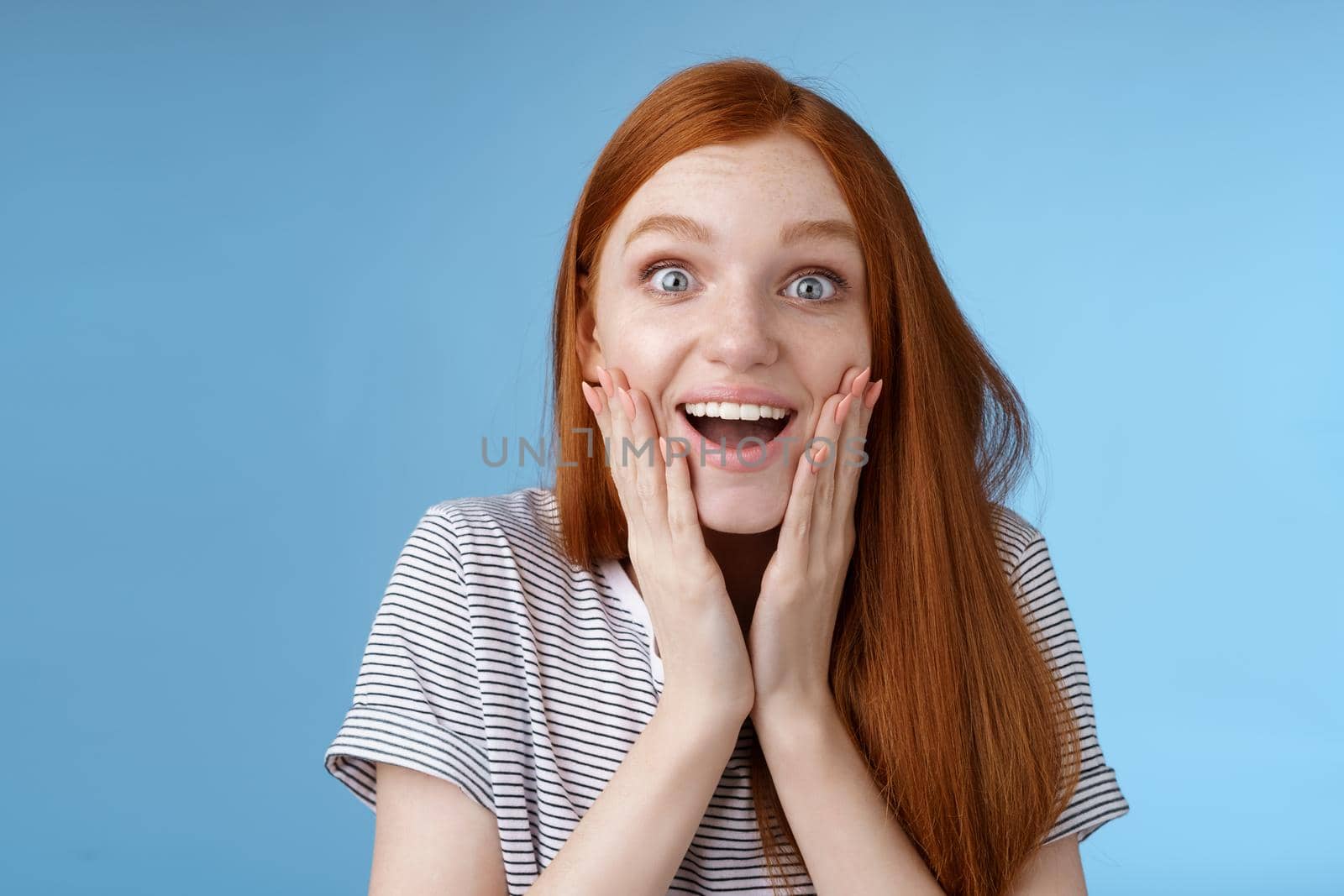 Amused happy dreamy young excited redhead female fan looking fascinated camera smiling admiration delighted touch face thrilled express emotional cheerful surprise, blue background.