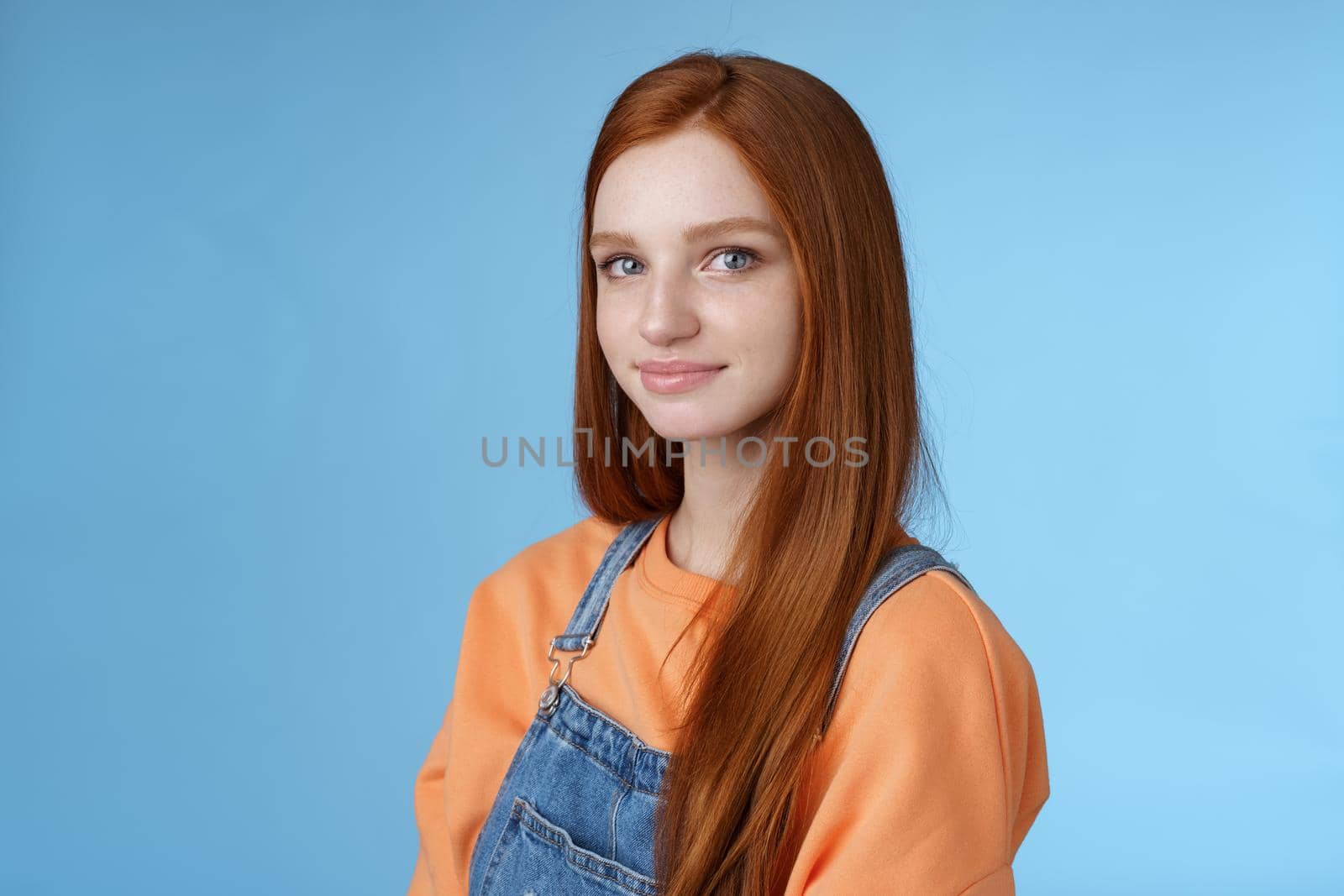 Waist-up kind sincere tender lovely redhead girl wearing orange shirt denim overalls standing half-turned smiling silly gentle grin camera looking friendly pleasantly walk alone blue background by Benzoix