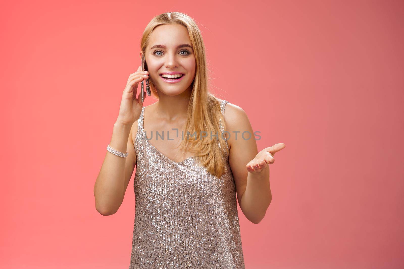 Joyful talkative outgoing attractive blond woman talking friend smartphone gesturing amused smiling broadly retelling fresh rumors after party wearing silver stylish dress, red background.