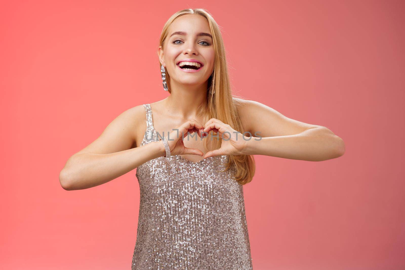 Adorable happy charming lovely blond european girl in silver dress express love positivity show heart gesture confess sympathy passionate feelings towards boyfriend smiling broadly, red background by Benzoix