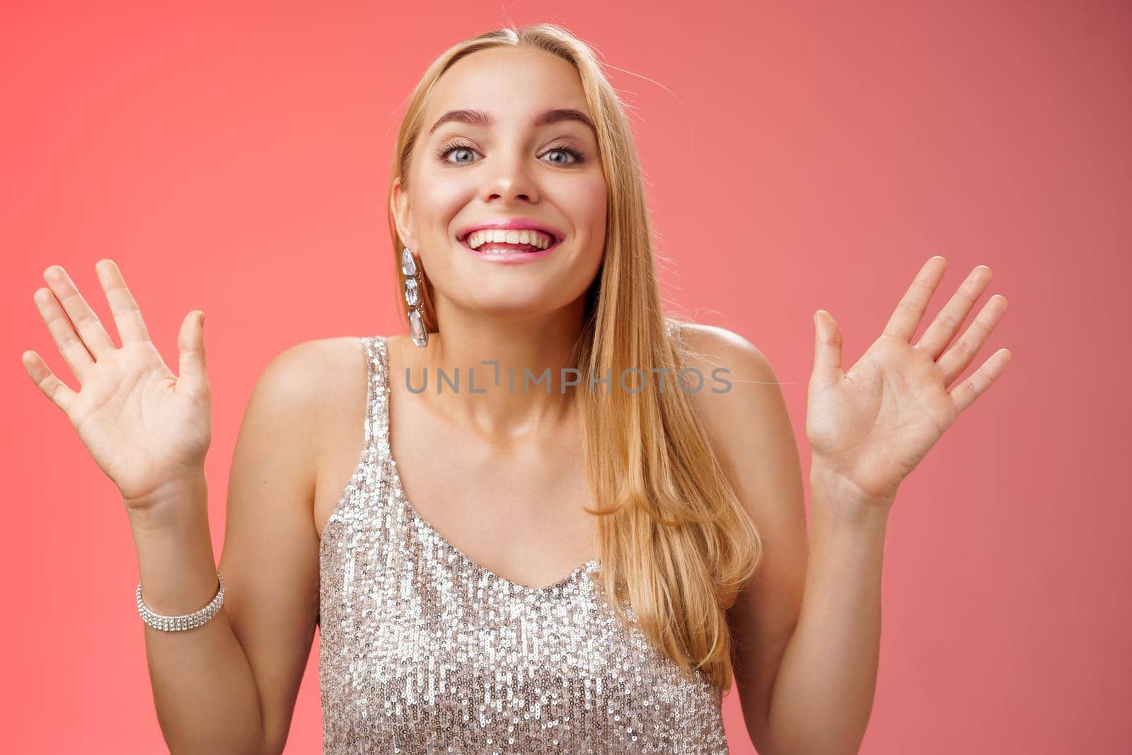 Happy elegant dreamy glamour young blond woman raising hands delight joyfully smiling camera glad see friends comming party welcoming guests grinning happily, wearing silver stylish dress.