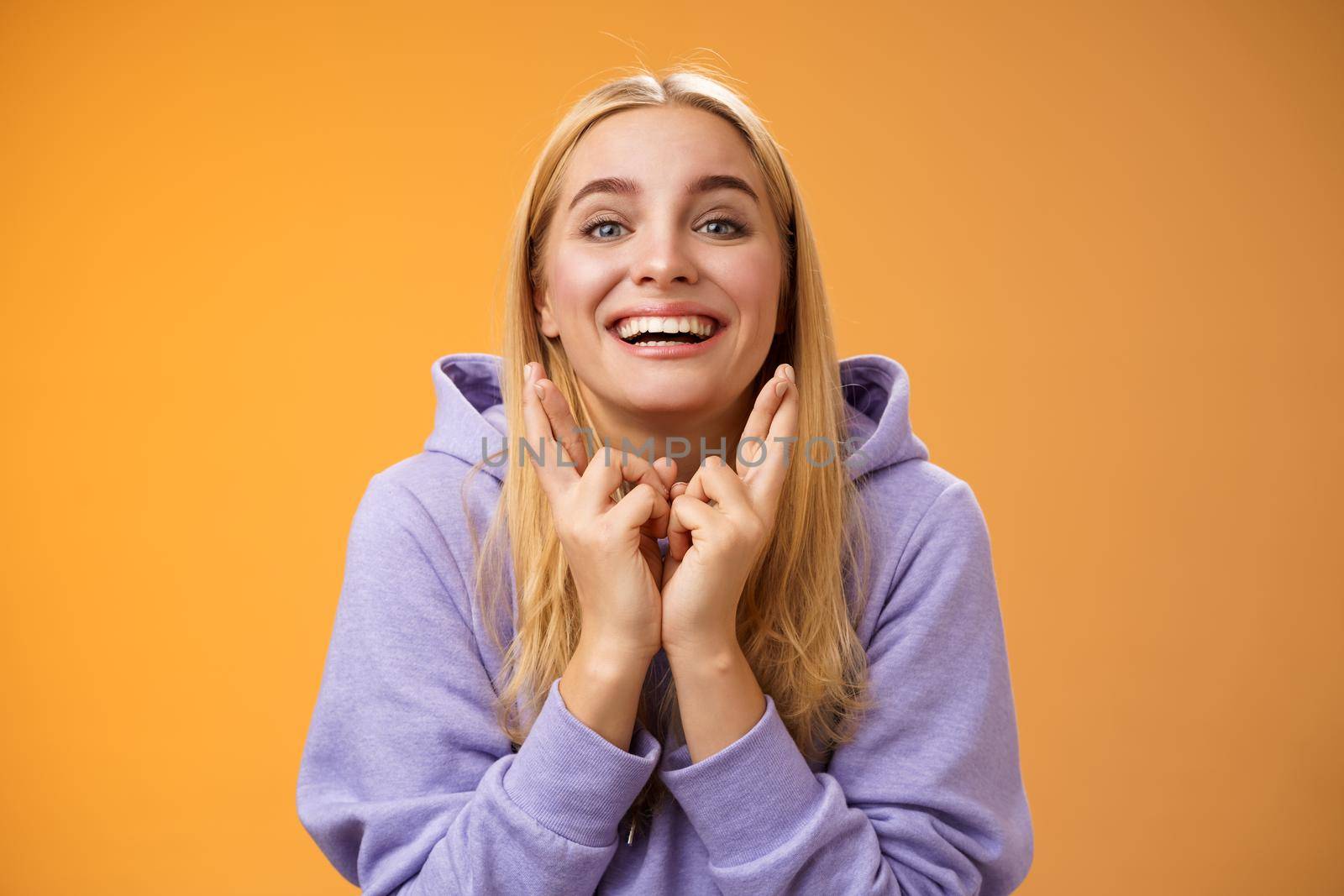 Lucky optimistic cute positive smiling blond girl grinning excited cross fingers making wish dreaming desire come true anticipating good news praying orange background waiting results by Benzoix