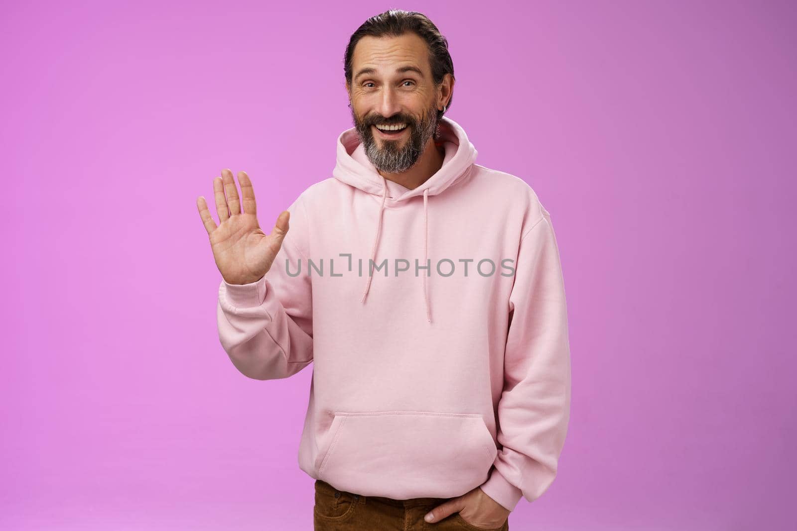 Cheeky charismatic funny happy smiling mature man bearded grey hair in pink hoodie waving palm hello nice meet you greetng gesture welcoming guests say hi standing purple background friendly relaxed by Benzoix