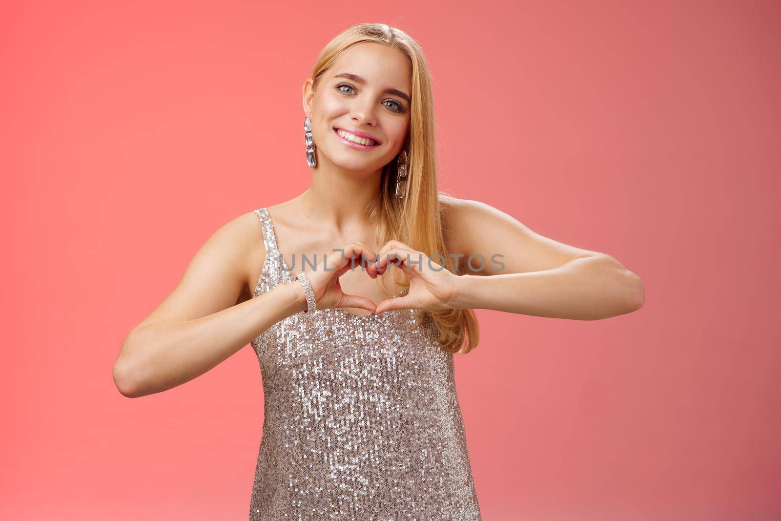 Romantic passionate charming lovely blond glamour woman in silver dress brilliants show heart love gesture express sympathy passion, adore relationship smiling delighted, standing red background.