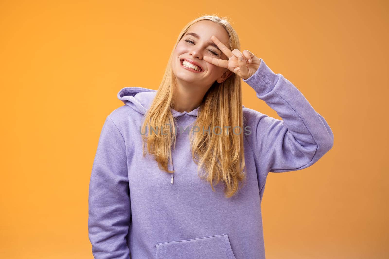 Carefree optimsitic young cute blond caucasian girlfriend having fun enjoying sun perfect spring weather winking cheeky smiling tilting head happily show peace victory sign, orange background.