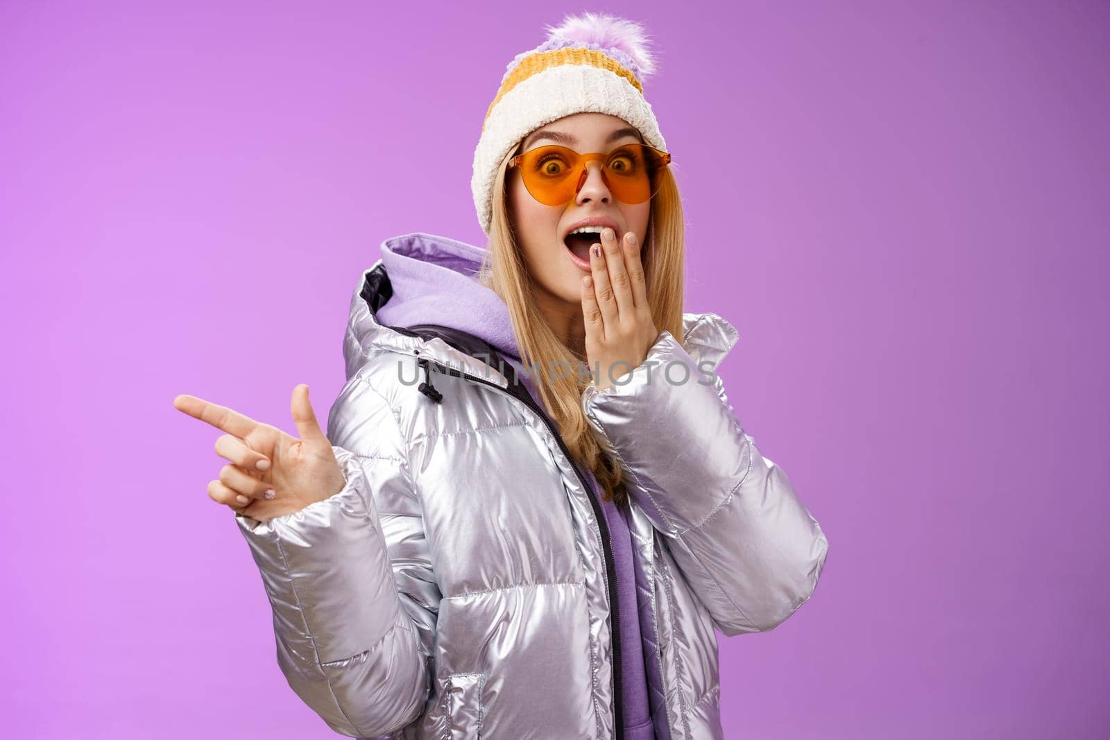 Amazed speechless surprised attractive stylish blond girlfriend in sunglasses, silver glittering jacket hat drop jaw impressed widen eyes shocked pointing left astonished, purple background by Benzoix